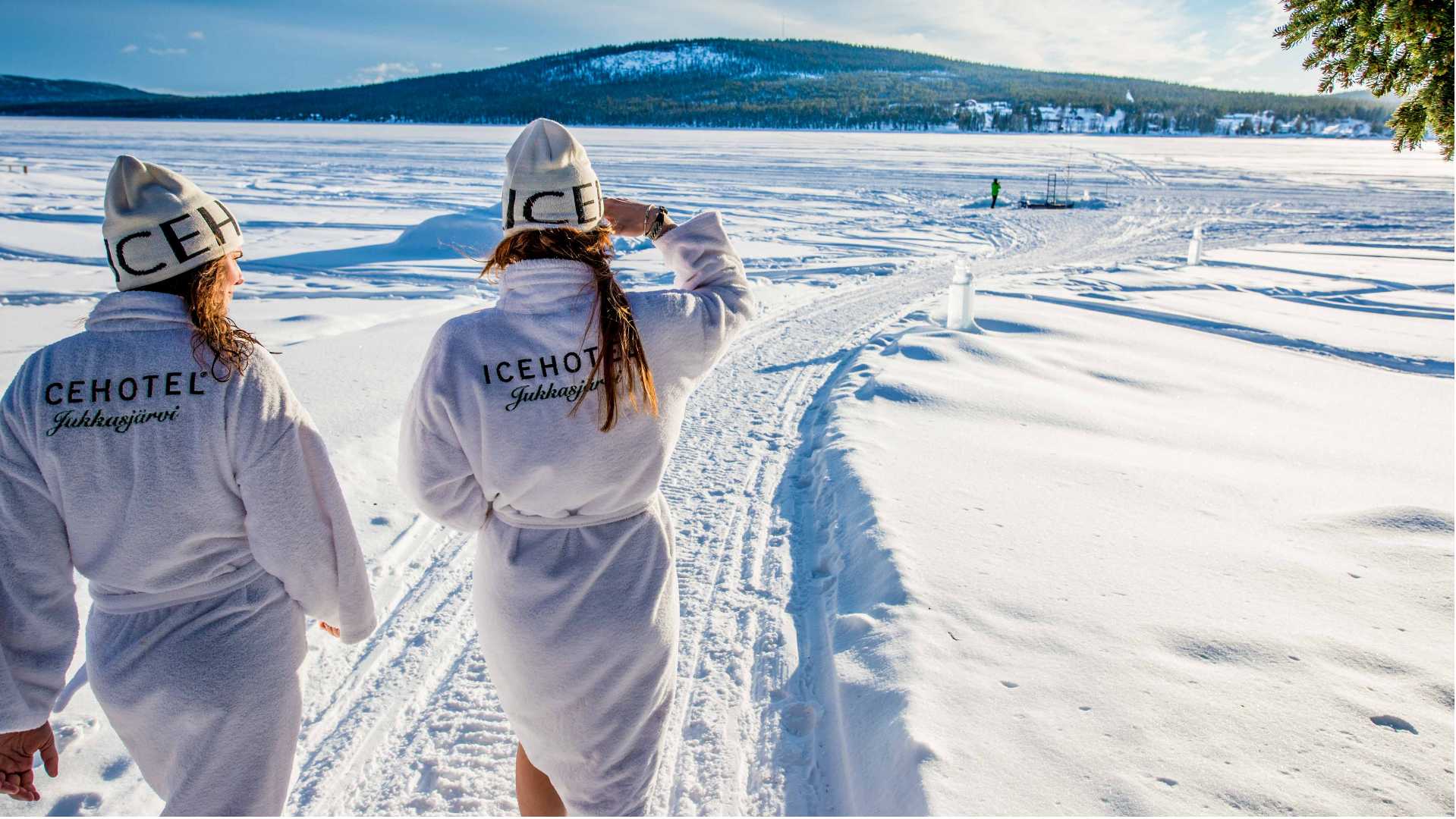 to kvinder i hvide badek&aring;ber og huer g&aring;r i sneen ved Icehotel i svensk lapland